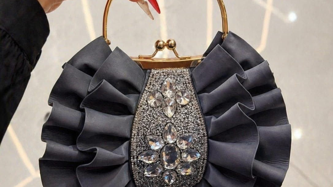 Handbag with ruffled design and gemstone details held by a hand against a light background