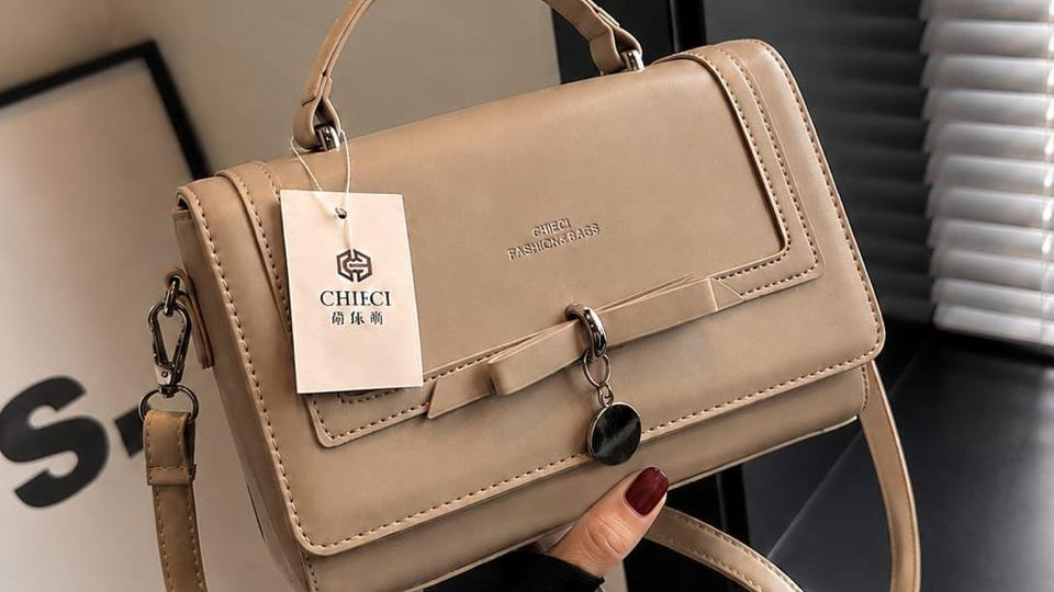 A beige top-handle handbag with a detachable shoulder strap, held by a hand. The bag features a front flap with a buckle detail, a hanging brand tag reading 'CHICCI', and a gold 'SHOP NOW' promotional badge overlay in the top-left corner.