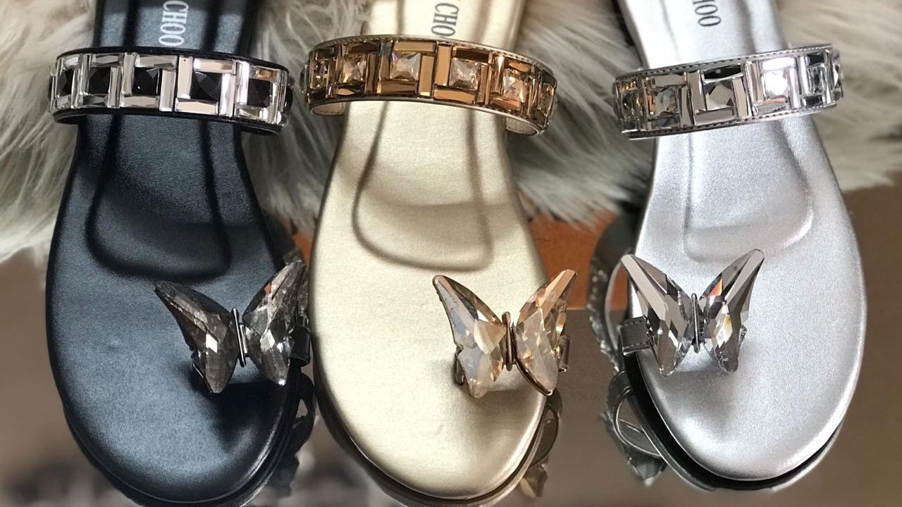 Three pairs of Jimmy Choo sandals with decorative straps and butterfly accents on a fluffy white surface.