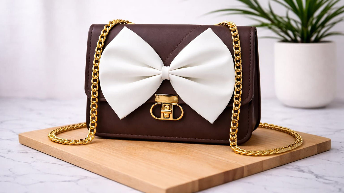Brown handbag with a large white bow and gold chain on a wooden board with a plant in the background.