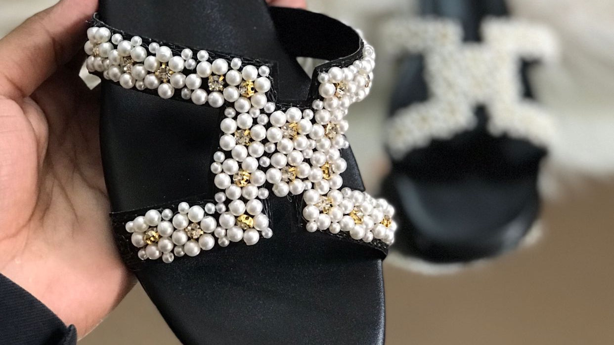 Black sandal with pearl embellishments held by a hand, Hermes logo visible.