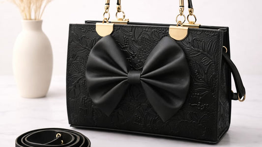 Black handbag with a large bow on a white surface, next to a vase with dried flowers.