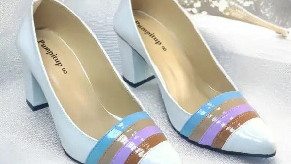 A pair of women's pointed-toe high heels in glossy white, featuring a decorative striped band across the vamp in light blue, lavender, and brown. The shoes are displayed on a white fabric background with a small dried flower prop.