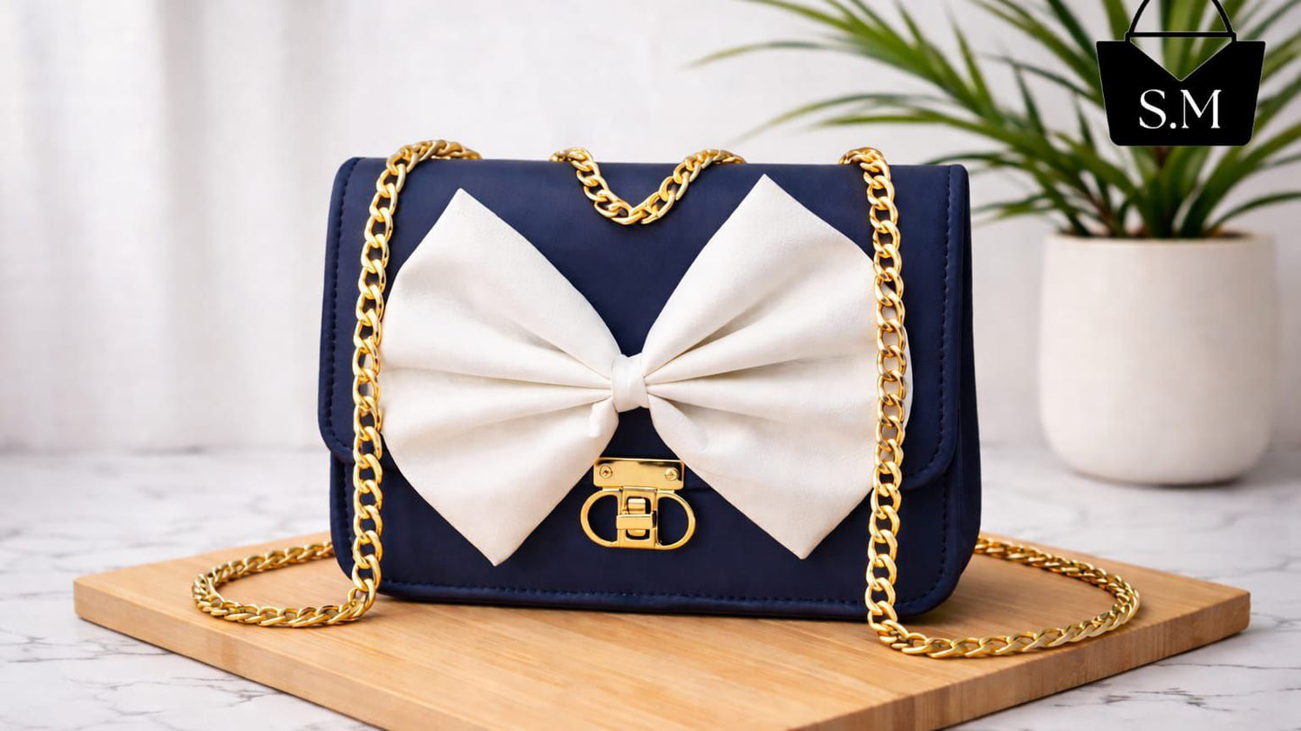 Navy blue handbag with a large white bow and gold chain strap on a wooden board.