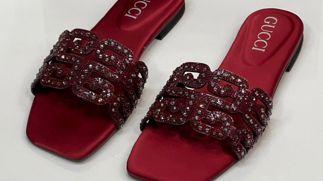Red Gucci sandals with embellishments on a white surface, Gucci box in the background.