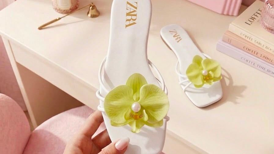 White sandals with green floral designs held by a hand in a pink room.