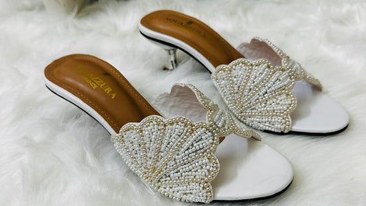 Pair of white sandals with beaded fan design on a white surface, Aquazzura branding visible.