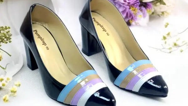 A pair of women's high-heeled pumps with a glossy black patent toe cap featuring diagonal stripes in light blue, lavender, beige, and white. The shoes are displayed on a draped white fabric surface with decorative flowers in the background.