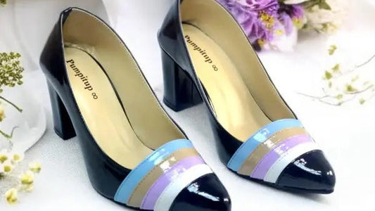 A pair of women's high-heeled pumps with a glossy black patent toe cap featuring diagonal stripes in light blue, lavender, beige, and white. The shoes are displayed on a draped white fabric surface with decorative flowers in the background.