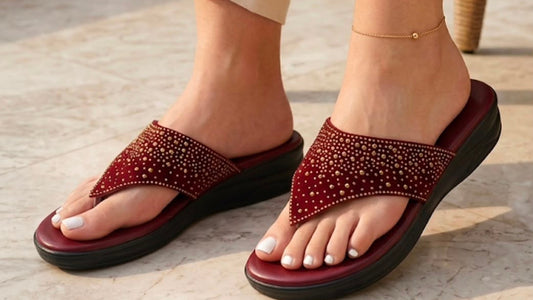 A person wearing burgundy slide sandals with sparkling rhinestone straps and beige linen pants, standing on a light stone floor next to a wicker chair.