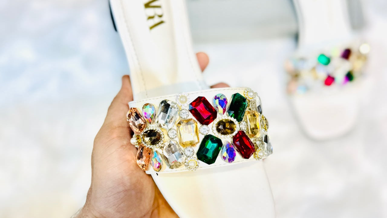 A hand holds a white slide sandal decorated with a large, colorful rhinestone and crystal embellished strap. Another matching sandal is visible in the background on a white textured surface.