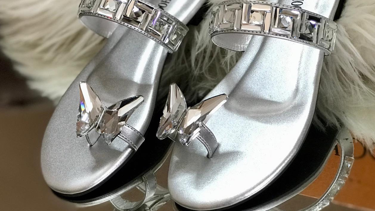 Silver sandals with gemstones on a fluffy white surface with 'Jimmy Choo' branding.