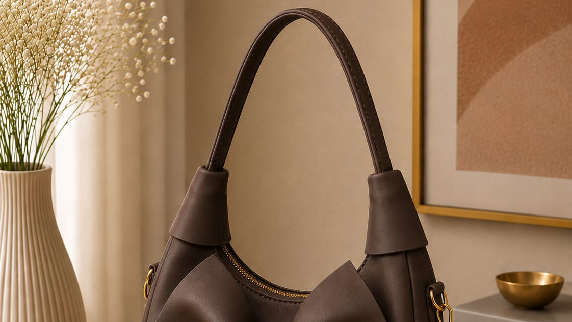 Brown handbag with a large bow on a beige surface, with a vase and books in the background.