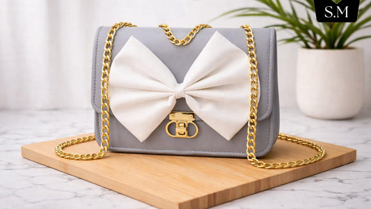 Gray handbag with a large white bow and gold chain on a wooden board, with a plant in the background.
