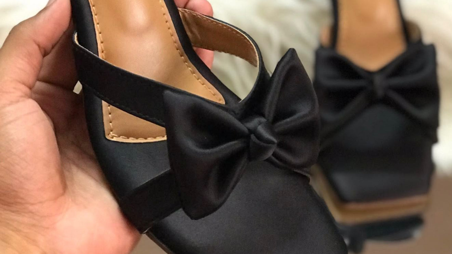 Black sandal with bow held by a hand, Jimmy Choo branding visible.