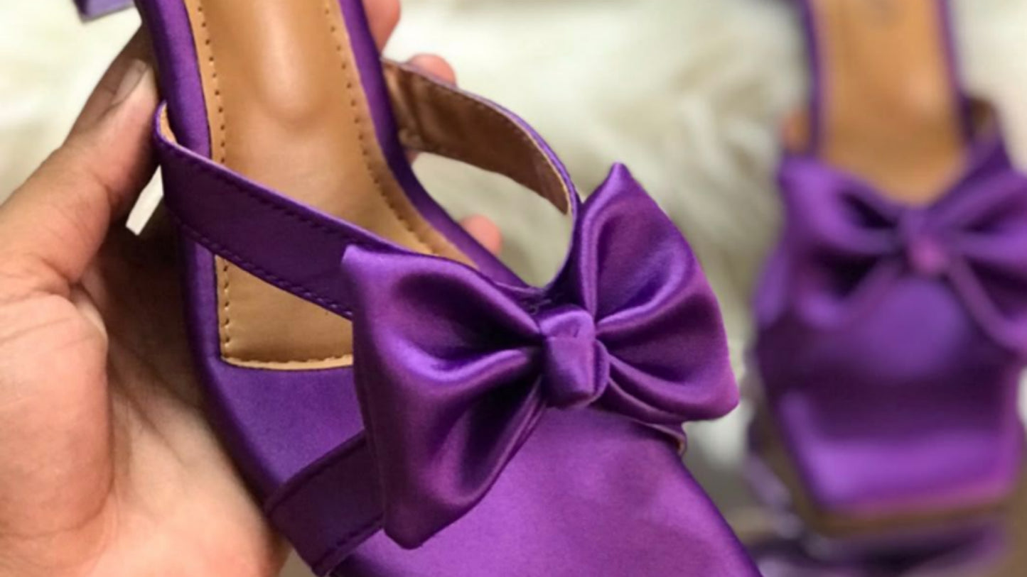Purple high-heeled shoes with a bow, featuring Jimmy Choo branding, held by a hand.