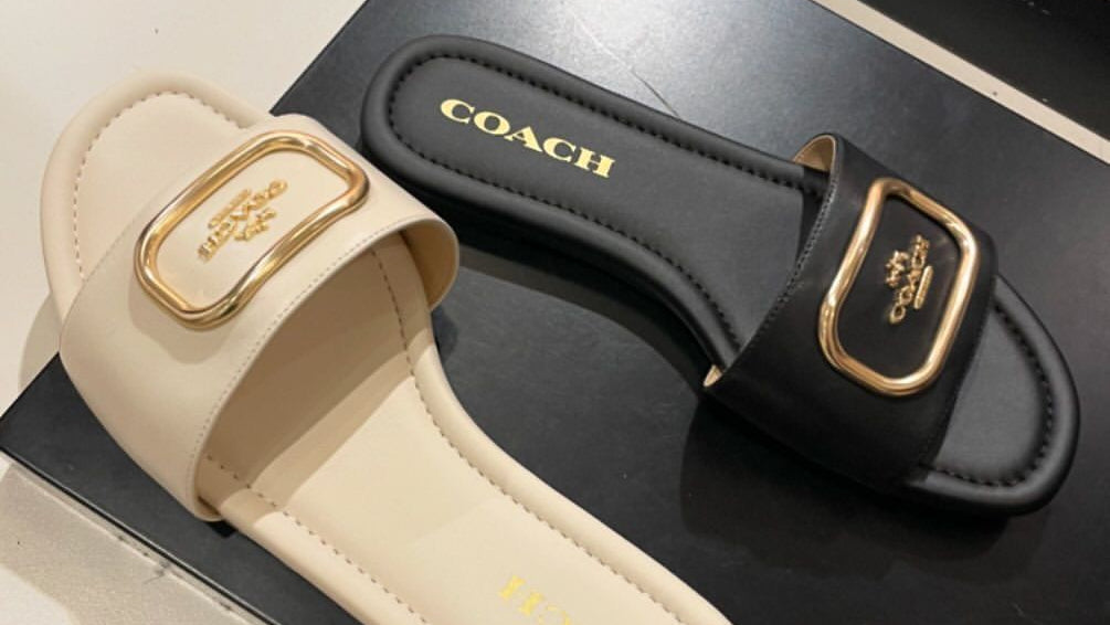 Beige and black Coach sandals with gold buckles on a black box.