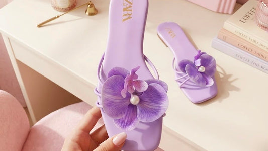 Purple sandals with floral design held by a hand in a room with a mirror and flowers.
