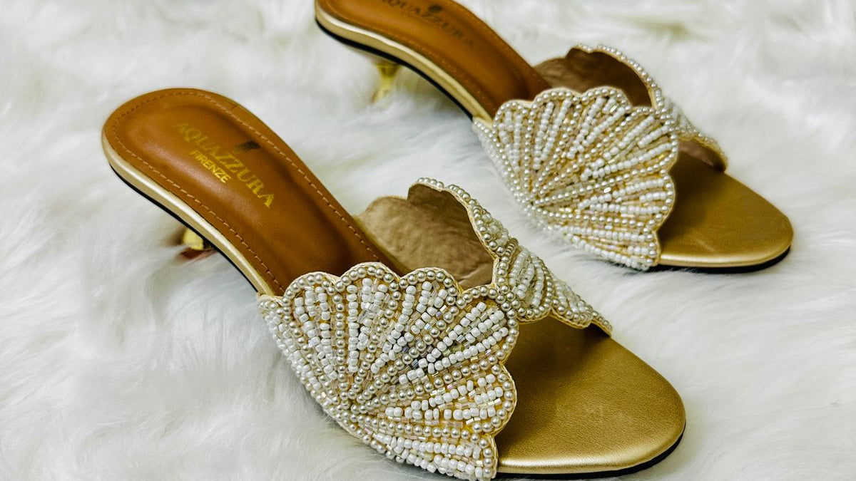 Gold high-heeled sandals with shell designs on a white fur surface, Aquazzura box in the background.