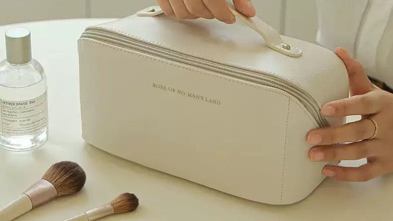 Person opening a beige cosmetic bag with makeup items on a light surface