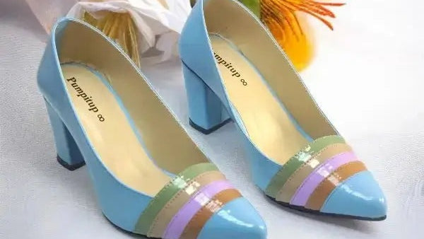 A pair of light blue high-heeled pumps with a pointed toe and a decorative multi-colored stripe detail in green, purple, and gold on the vamp. The shoes are displayed on a white fabric background with decorative white and orange flowers.