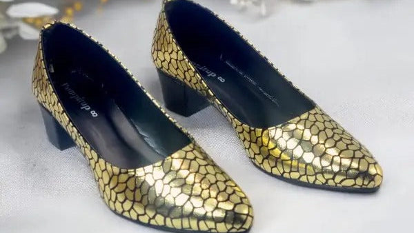Pair of gold mosaic high heels on a white surface with flowers in the background