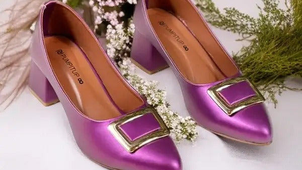 Purple high-heeled shoes with gold buckles on a white surface with flowers in the background