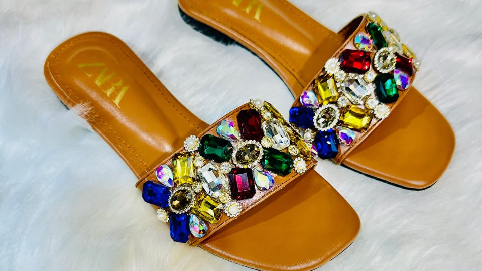 A pair of tan leather slide sandals with wide straps heavily embellished with colorful faux rhinestones in red, green, blue, yellow, and purple, displayed on a white faux fur background.