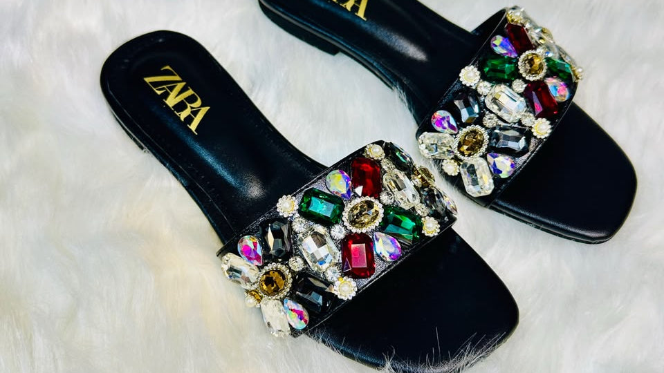 A pair of black Zara slide sandals embellished with colorful rhinestones, displayed on a white faux fur surface with a Zara shoebox in the background.