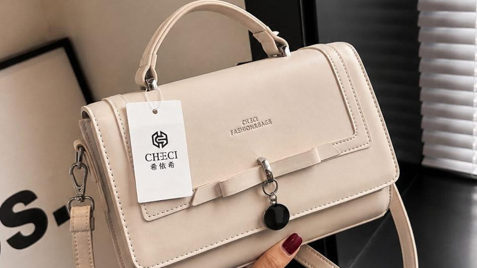 A light beige structured handbag with a top handle, shoulder strap, and front flap, held by a hand. The bag features a small bow detail on the front, a hanging black charm, and a white brand tag reading 'CHEKI'.