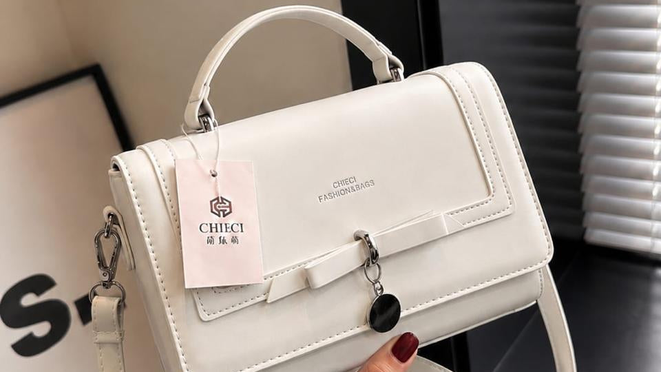 A white structured handbag with a top handle, front flap, and long shoulder strap, held by a hand. The bag has a pink hangtag with the brand name CHICLE.